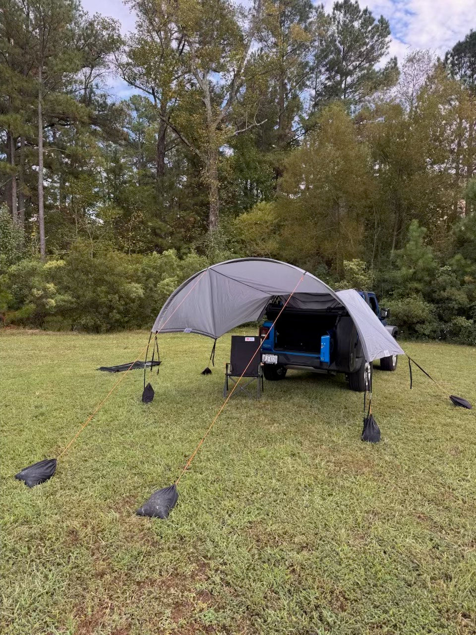 Trail Gear Oasis 2-in-1 cover deployed in shade mode off the rear of a blue Jeep Wrangler JL 2-door, staked out on grass with trees in the background