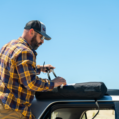 Man in plaid shirt setting up Trail Gear Oasis SUV sun shade from roof-mounted storage bag