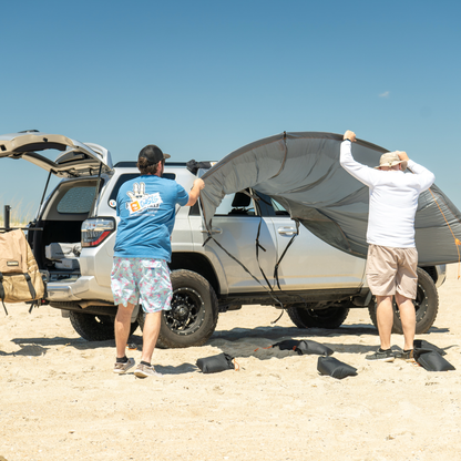 Two people deploying Trail Gear Oasis SUV trail cover sun shade on beach with sandbag anchors