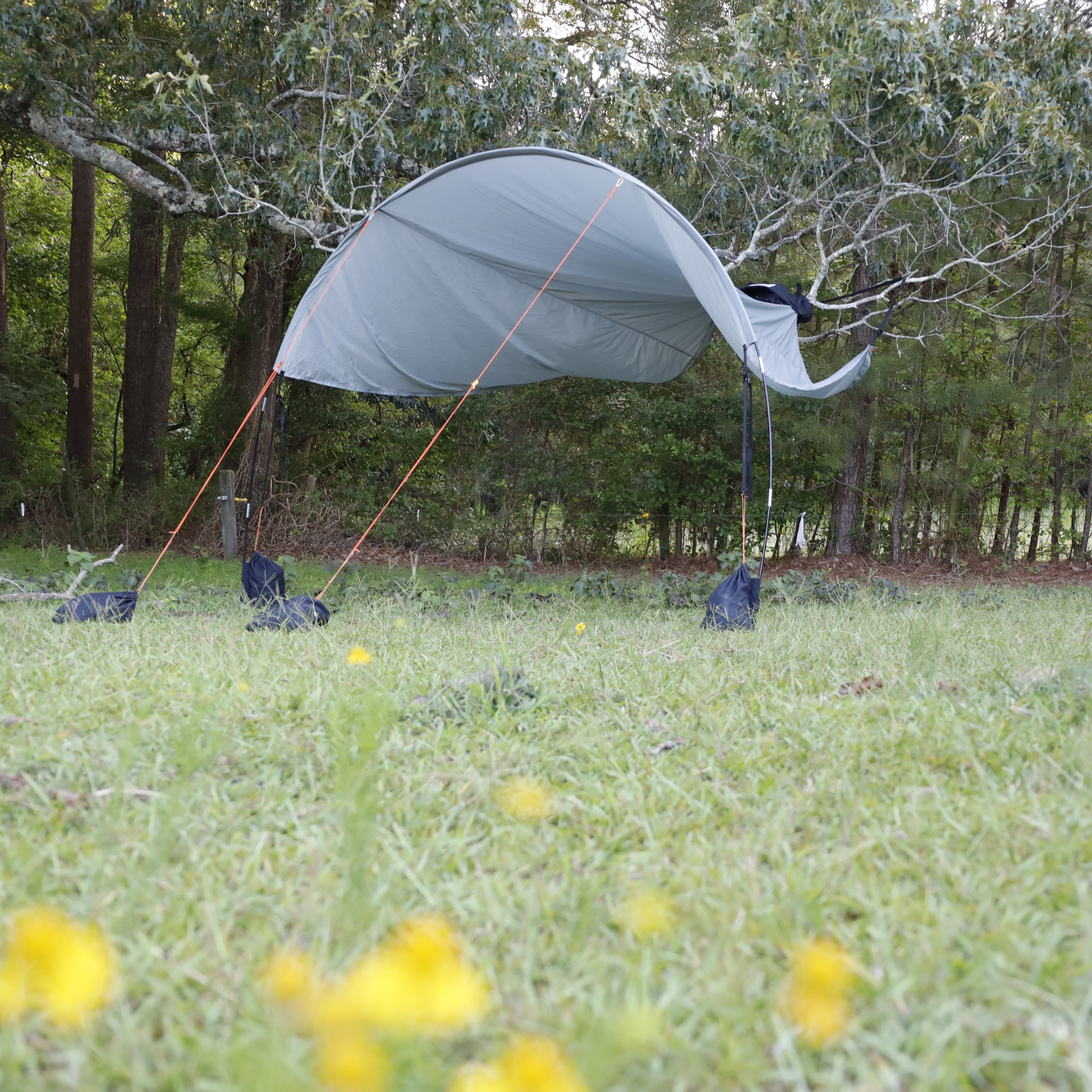 Trail Gear Oasis Sidekick Shade suspended between trees with orange guy lines and sandbag anchors in wooded area